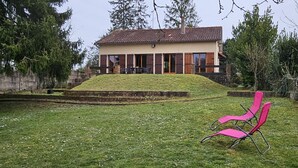 Una casa con techo inclinado, un porche con sillas y un jardín cubierto de césped.