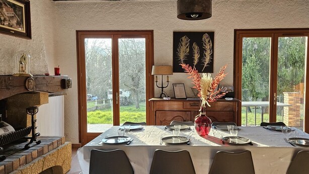 Comedor con una mesa preparada para cuatro personas, un florero rojo con flores secas y vista a la vegetación exterior.