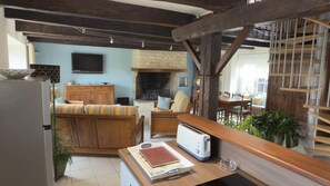 A rustic living room with exposed beams, a fireplace, a dining area, and a kitchen with a toaster. - Au bord du Clain - ROMAGNE (ROMAGNE)