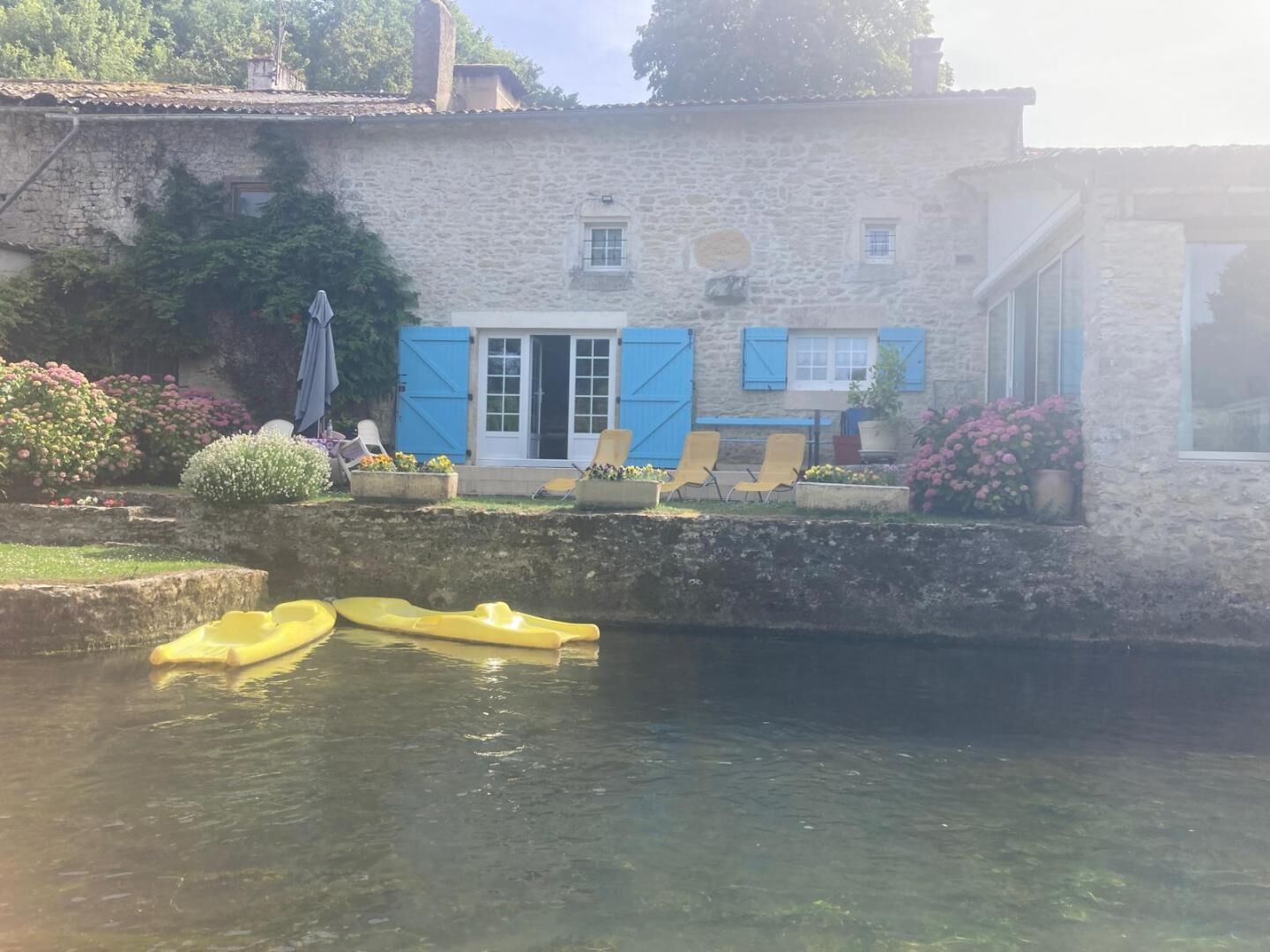 Un bâtiment en pierre avec des volets bleus, un petit jardin fleuri et un canal où flottent des bateaux jaunes.