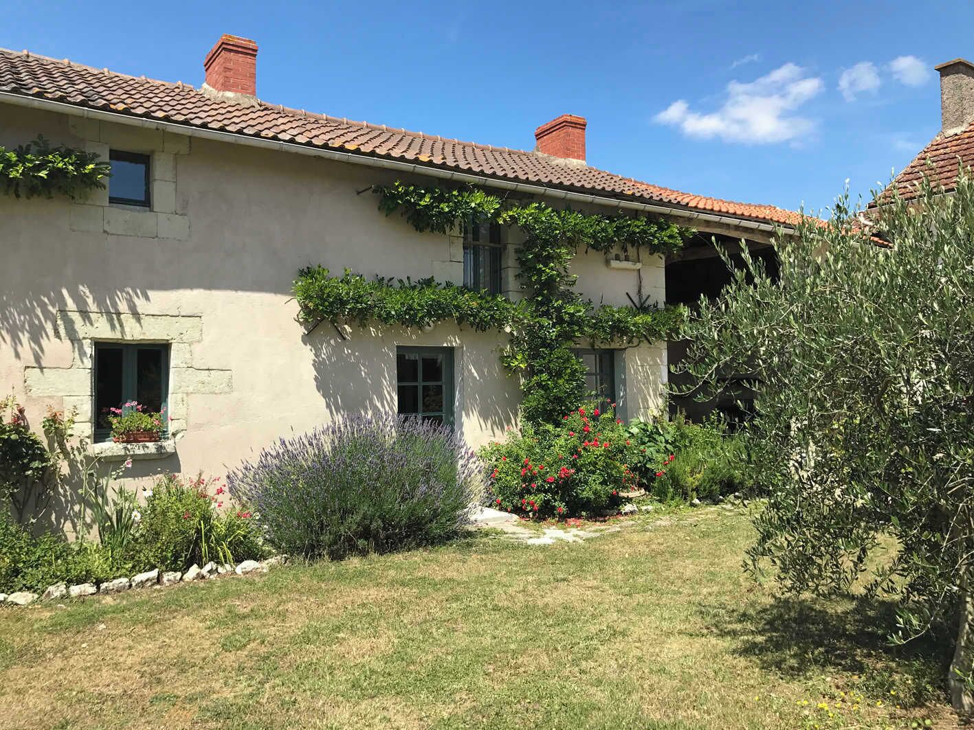 Une maison avec un toit en tuiles, une petite fenêtre agrémentée d’une jardinière et un jardin où poussent différentes plantes et fleurs.