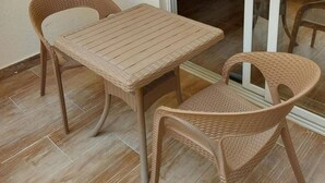 Een balkon met een tafel en twee rieten stoelen, een televisie op de achtergrond en een schuifdeur met glas.