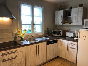 A kitchen with wooden cabinets, a built-in oven, a sink, and a window with curtains. - La Petite Maison Bleue (SOSSAIS)