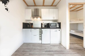 A compact kitchen with white cabinets, a stainless steel range hood, a microwave, and a dishwasher. There is a bunk bed visible through an open doorway. - Le Gite du Prunus - MONTAMISE (MONTAMISE)