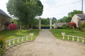 Un chemin de gravier mène à une entrée fermée par un portail, avec un bâtiment en pierre sur la gauche et un mur en pierre sur la droite.