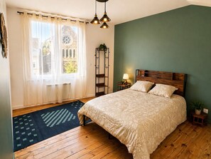 Une chambre à coucher avec un lit, une fenêtre avec des rideaux, un radiateur, un plancher en bois et un luminaire suspendu.