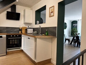 Une cuisine moderne équipée d’un four, d’un micro-ondes et d’un évier encastrés. On aperçoit un coin repas avec une table et des chaises à travers une porte ouverte.