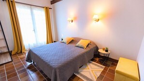 Ein Schlafzimmer mit einem Bett, zwei Nachttischlampen, einem kleinen Tisch mit einer Pflanze, einem gelben Hocker und einem gemusterten Teppich.