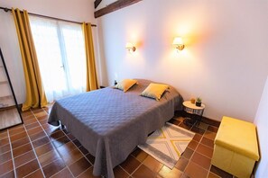 Ein Schlafzimmer mit einem Bett, zwei Nachttischlampen, einem kleinen Tisch mit einer Pflanze, einem gelben Hocker und einem gemusterten Teppich.