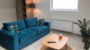 En moderne stue med en blå sofa, et lille træbord, en fjernbetjening, en potteplante og et vindue med udsigt.