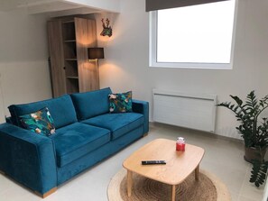 A modern living room with a blue sofa, a small wooden table, a remote control, a potted plant, and a window with a view. - Le Loft 2 - VOUNEUIL SOUS BIARD (VOUNEUIL SOUS BIARD)