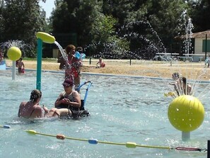Ein Schwimmbecken mit Wasserstrahlmassagen und treibenden Bällen, umgeben von einer Sandfläche mit Bäumen.
