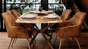 Una mesa de comedor con sillas, una lámpara colgante y vistas a árboles a través de amplios ventanales.