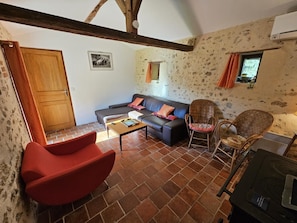 A living room with a sofa, a red chair, a wicker chair, a table, and a wooden ceiling beam. - Gîte de la Vacherie - VIGEANT (LE) (VIGEANT (LE))