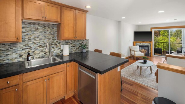 A modern kitchen with wooden cabinets, a black countertop, and a dishwasher. There is a dining area with a table and chairs, and a fireplace. - Modern City Retreat • Family Friendly • Free Prkg (Seattle)