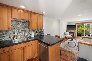 A modern kitchen with wooden cabinets, a black countertop, and a dishwasher. There is a dining area with a table and chairs, and a fireplace. - Modern City Retreat • Family Friendly • Free Prkg (Seattle)