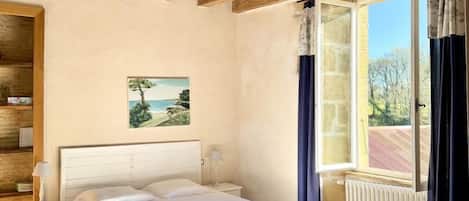 A bedroom with a wooden ceiling, a bed, a nightstand, a table, a window with curtains, and a painting on the wall.