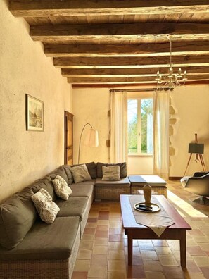 A cozy living room with a sectional sofa, a wooden table, and a large window with curtains. - The House of Villars (PRESSAC)