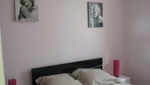 Un dormitorio con una cama, dos almohadas, una mesita de noche con un objeto rojo y dos cuadros colgados en la pared.