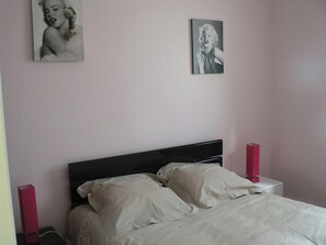 Ein Schlafzimmer mit einem Bett, zwei Kissen, einem Nachttisch mit einem roten Gegenstand und zwei Bildern an der Wand.