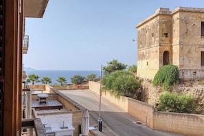 A view from a balcony overlooking a coastal road with a historic stone building and a clear view of the sea.
