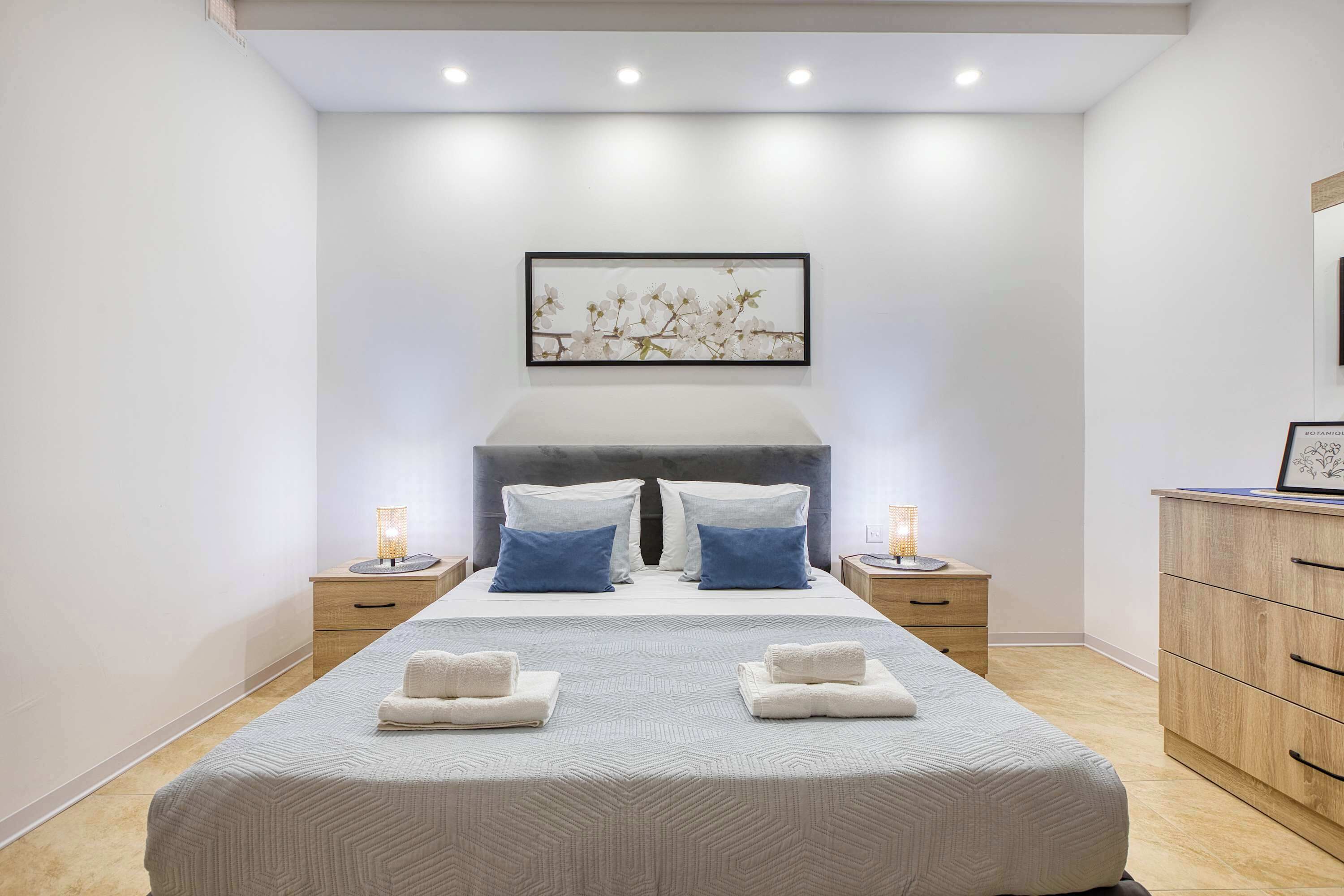 A modern bedroom with a large bed, two nightstands, a dresser, and a wall-mounted artwork.