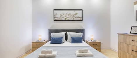 A modern bedroom with a large bed, two nightstands, a dresser, and a wall-mounted artwork.