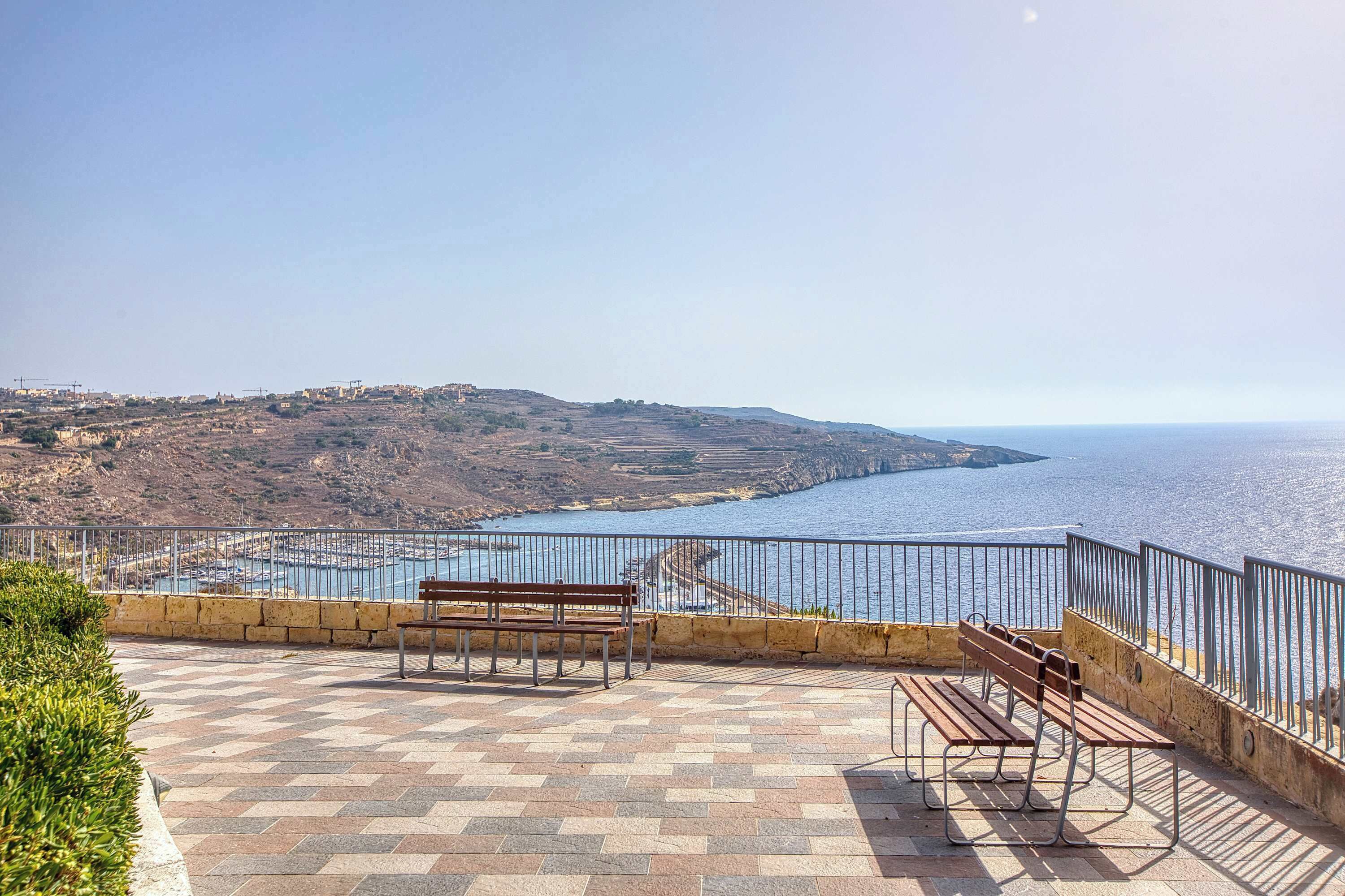 Ein Aussichtspunkt an der Küste mit einer gepflasterten Terrasse, Bänken und einem Geländer mit Blick auf das Meer und die entfernten Hügel.