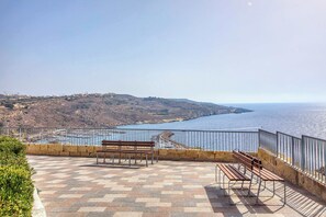 Un point de vue panoramique sur la côte, doté d’une terrasse dallée, de bancs et d’une balustrade offrant une vue sur la mer et les collines lointaines.