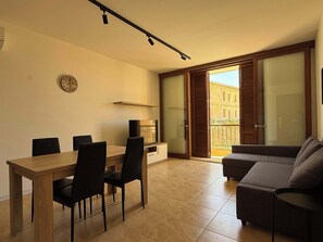 A modern living room with a dining area, a TV, a clock, and a sliding glass door to a balcony.