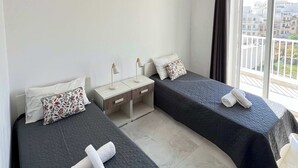 Habitación de hotel con dos camas, una mesita de noche, ventilador de techo y un ventanal con vistas a edificios.