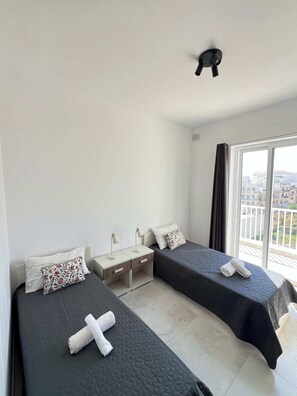 Ein Hotelzimmer mit zwei Betten, einem Nachttisch, einem Deckenventilator und einem großen Fenster mit Blick auf Gebäude.