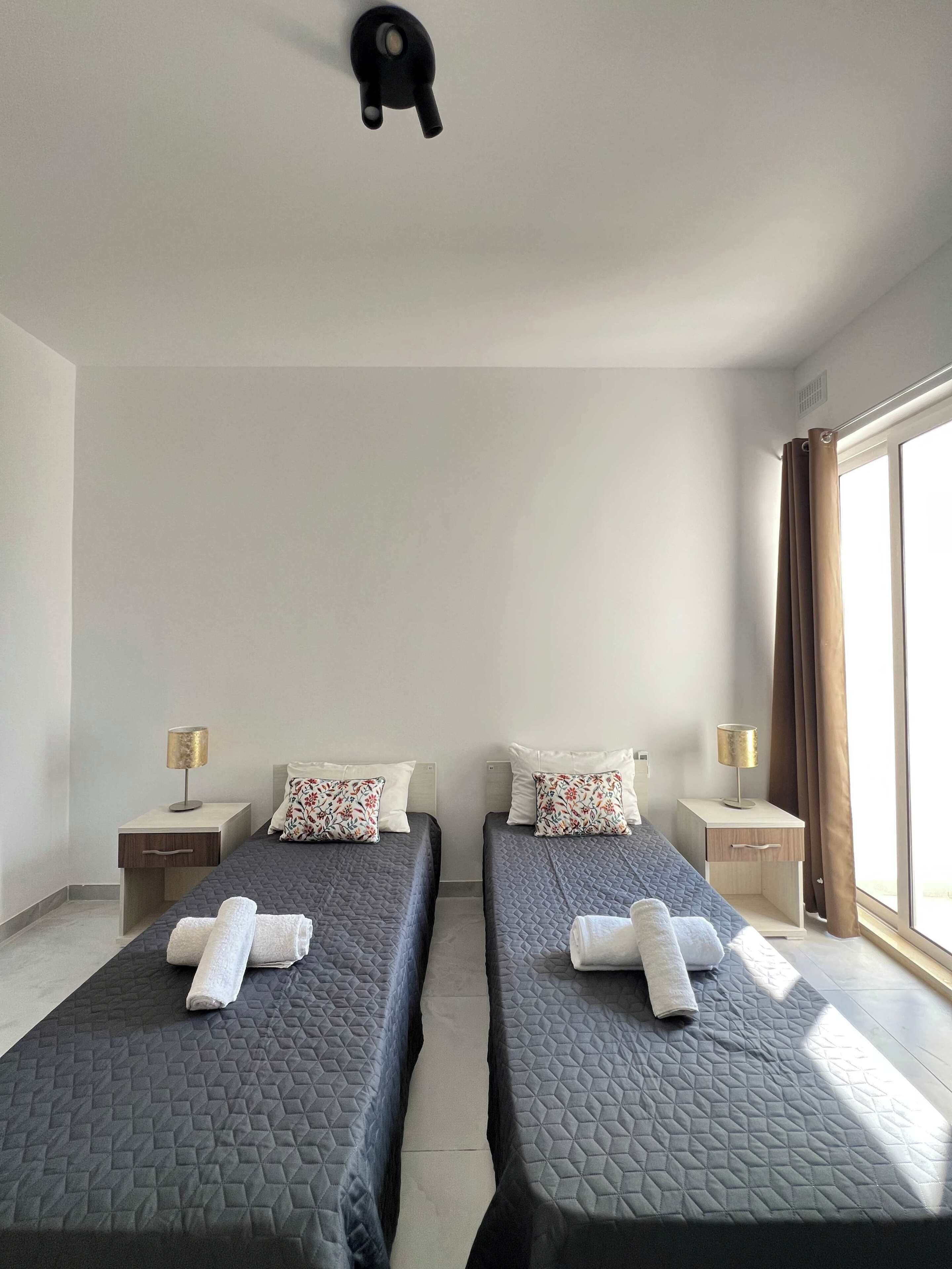 A hotel room with two beds, each with a quilted mattress and a pillow. There are bedside tables with lamps and a window with curtains.