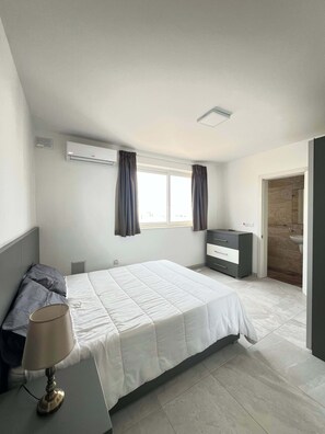 A bedroom with a bed, a dresser, an air conditioning unit, a radiator, and a bathroom visible through an open door. - Swieqi Spacious 3 BDR Apartment by PMI Malta (Is-Swieqi)
