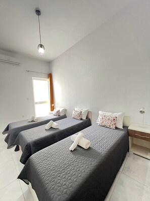 A hotel room with two beds, a nightstand, a lamp, and a window with curtains. - Swieqi Large 2BDR Maisonette with Yard by PMI (Swieqi)