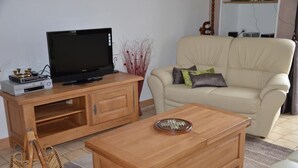 Una sala de estar con un televisor sobre un mueble de madera, un sofá color beige con cojines, una mesa de centro de madera y una cesta de mimbre.