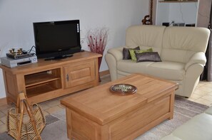 A living room with a television on a wooden stand, a beige sofa with cushions, a wooden coffee table, and a wicker basket. - Les Lilas - L'HOMMAIZE (LHOMMAIZE)
