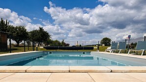 Una piscina rectangular con un patio empedrado al frente, rodeada por una cerca y árboles.