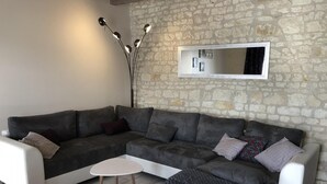 A living room with a grey sectional sofa, a small white table, and a stone wall. - The 3 Little Pigs (ROCHE-RIGAULT (LA))