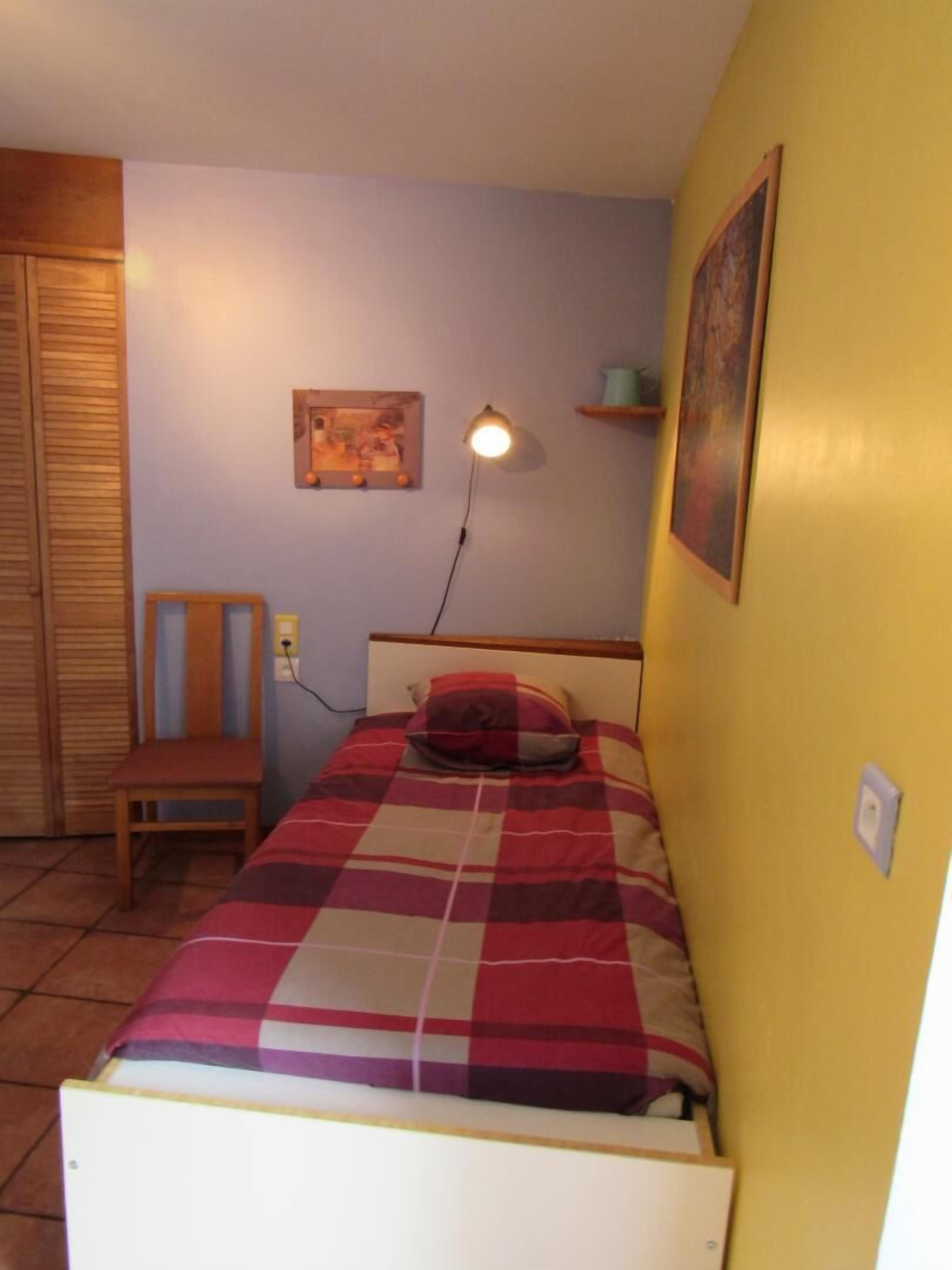 A small, single-bed room with a wooden headboard, a bedside lamp, a chair, and a wall-mounted shelf.