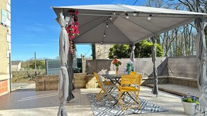 Een overdekt terras buiten met een tafel en stoelen, een patroonrijk kleed en decoratieve bloemen.