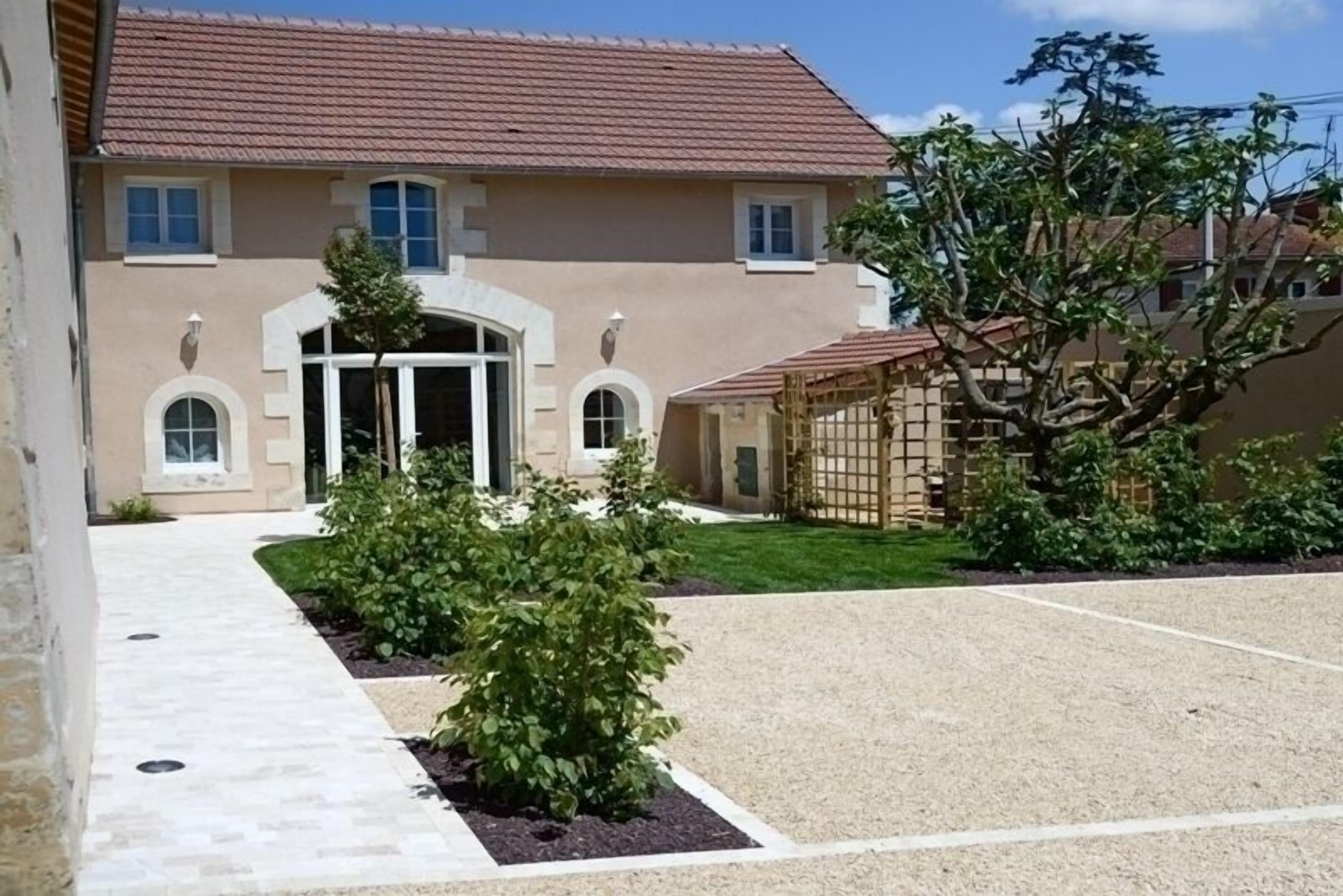 Un bâtiment de couleur beige, avec un toit en tuiles, une entrée centrale et une allée gravillonnée.