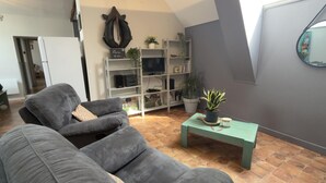 Ein Wohnzimmer mit grauem Sofa, einem grünen Couchtisch, einem Fernseher auf einem weißen Lowboard und einem Bücherregal mit Pflanzen.