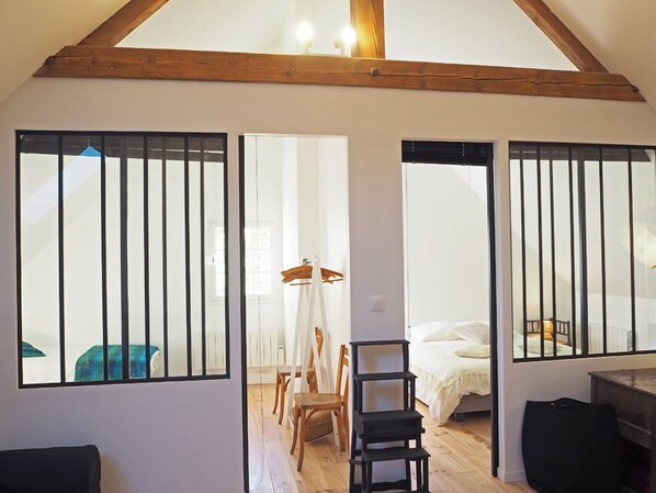 Une pièce avec un plafond en bois, un lit avec une literie blanche, une chaise, une petite table et une fenêtre avec des stores verticaux.