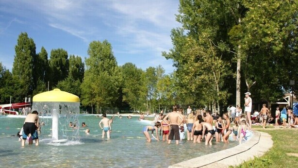 Piscina com pessoas, uma fonte de água e árvores ao fundo.