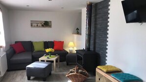 Een woonkamer met een bank, poef, televisie, open haard en een aan de muur bevestigde televisie.