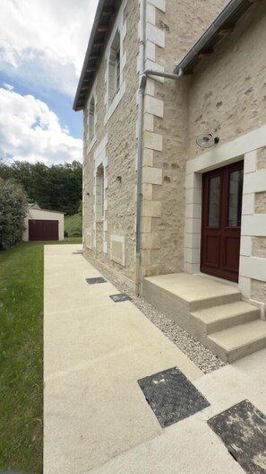 A stone building with a red door, a concrete pathway, and a garage in the background. - Au clair de la Rhune (MARCAY)