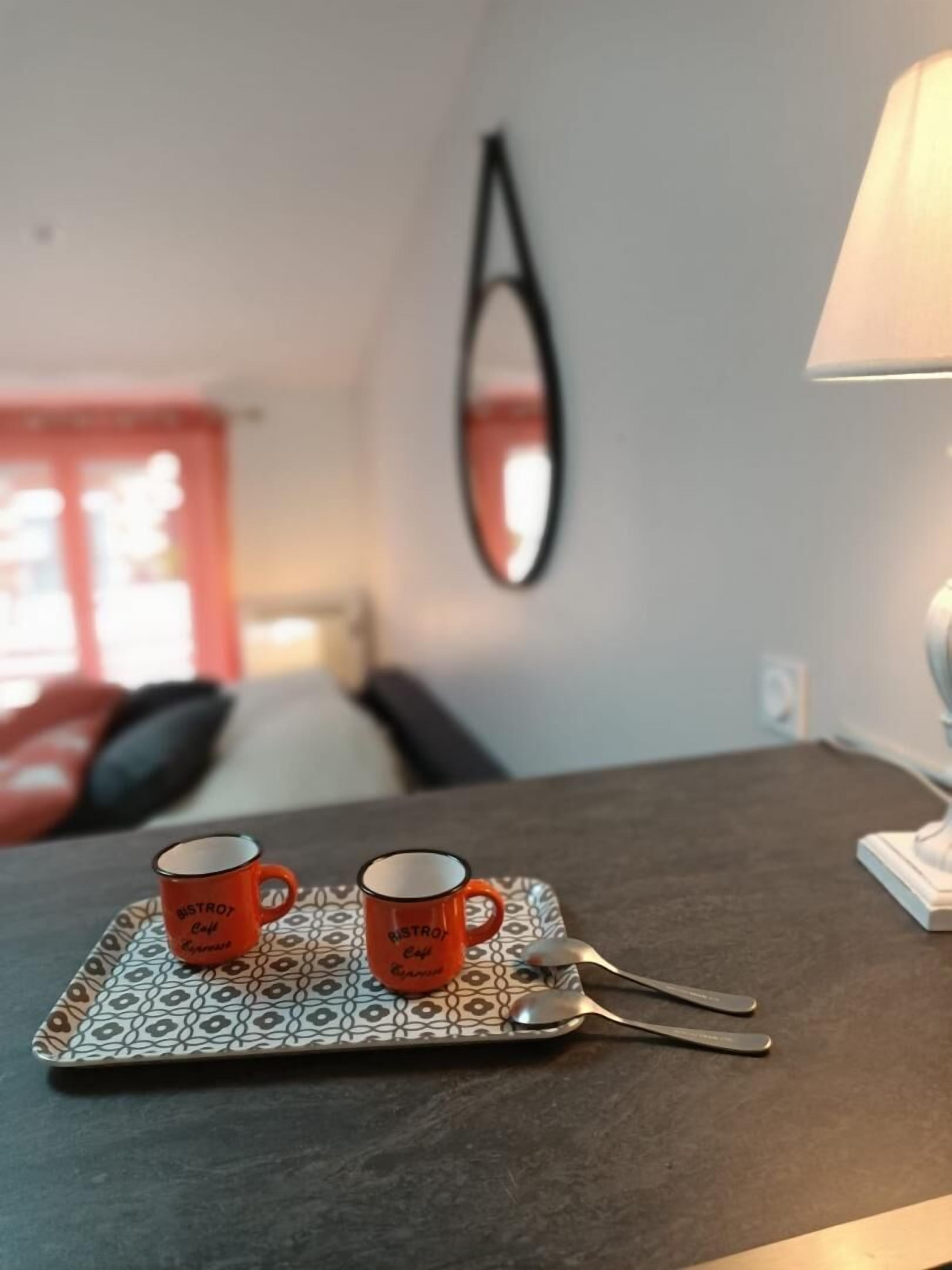 A table with two mugs on a tray, a spoon, and a lamp on a table in a room with a mirror on the wall.