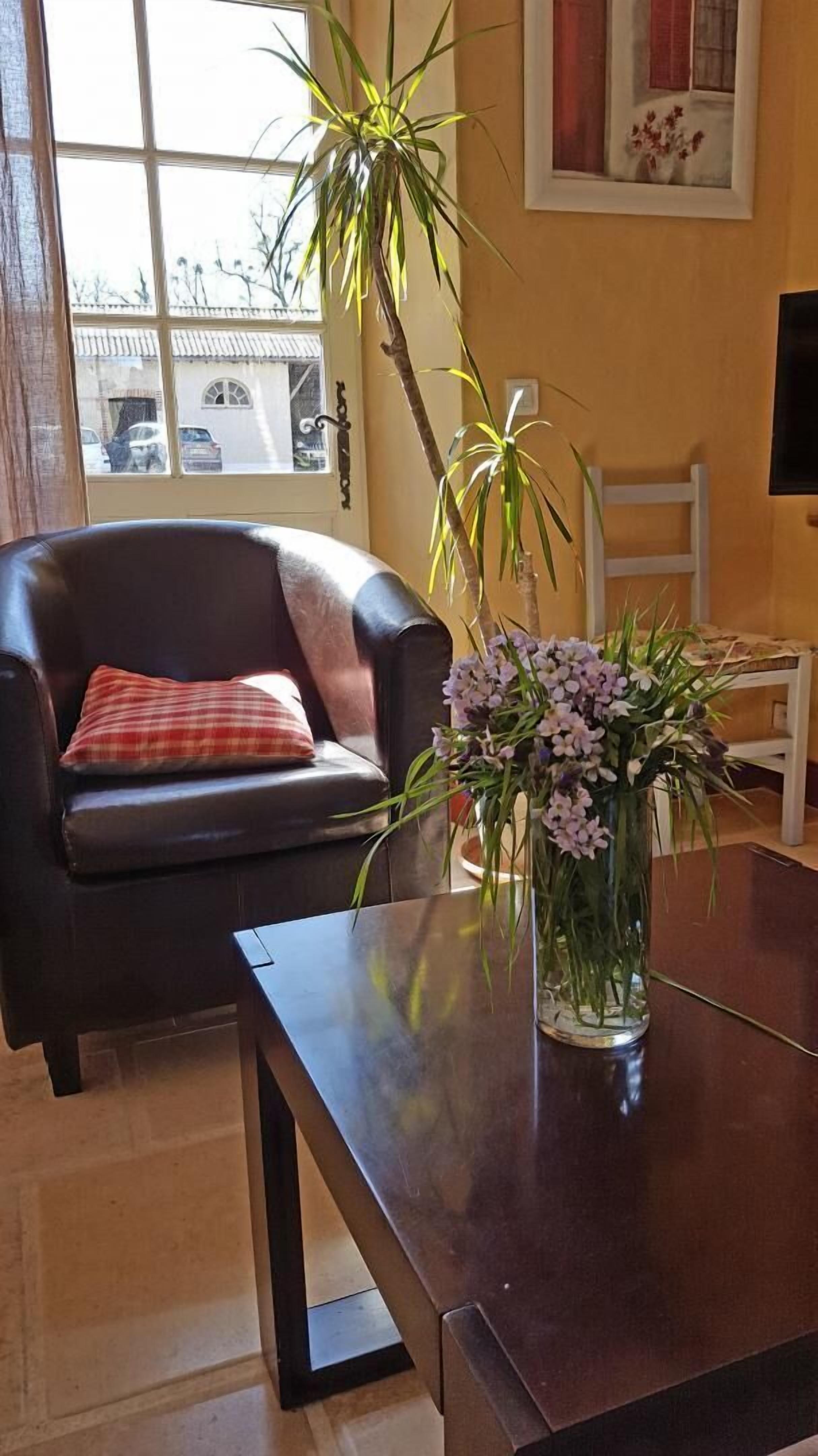 Ett rum med en fåtölj i läder, ett träbord, en vas med blommor och ett fönster med utsikt över byggnader.