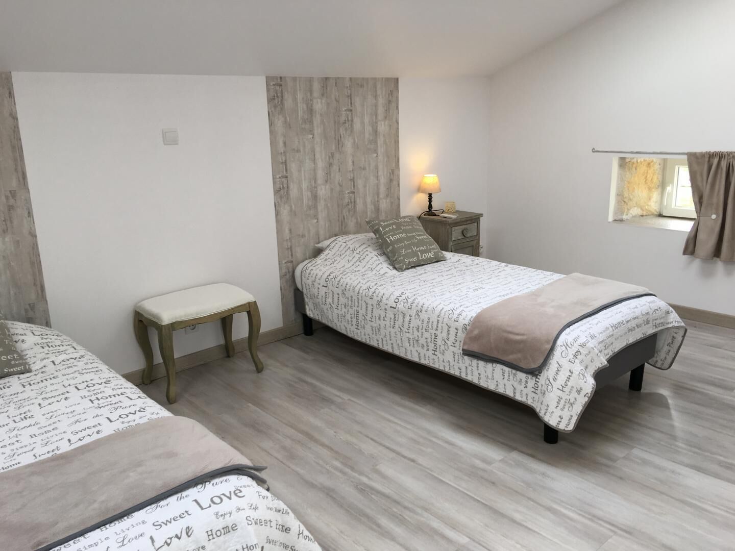 Un dormitorio con dos camas, una mesita de noche con lámpara, una ventana con cortinas y un banco de madera.
